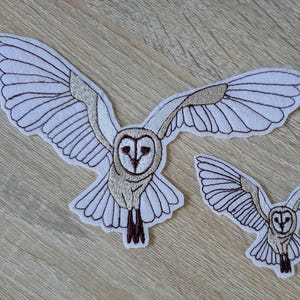 May include: Two embroidered owl patches with outstretched wings. The owls have white bodies, detailed brown wing outlines, and tan and brown facial features. One patch is larger than the other, both are on a light wood grain surface.