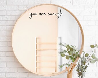 May include: A round wooden framed mirror with the text "you are enough" written in black cursive on the mirror surface. The mirror is hanging on a white brick wall.