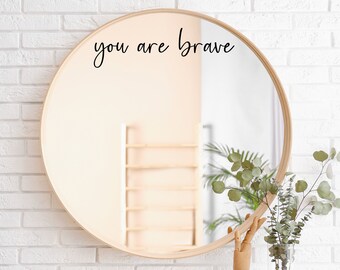 May include: A round wooden framed mirror with the text "you are brave" written in black cursive on the mirror's surface. The mirror is hanging on a white brick wall.