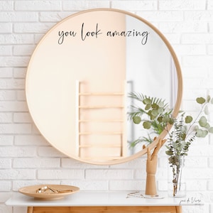 May include: A round wooden framed mirror with the text "you look amazing" written in black cursive on the mirror surface. The mirror is hanging on a white brick wall.