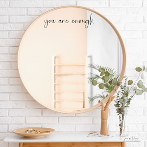 May include: A round wooden framed mirror with the text "you are enough" written in black cursive on the mirror surface. The mirror is hanging on a white brick wall.