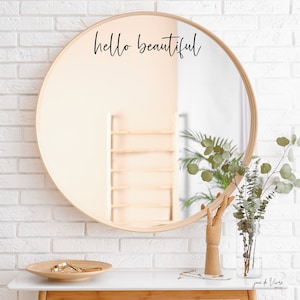 May include: A round wooden framed mirror with the text "hello beautiful" written in black script on the mirror surface. The mirror is hanging on a white brick wall. A vase with green eucalyptus branches is on a white table in front of the mirror.