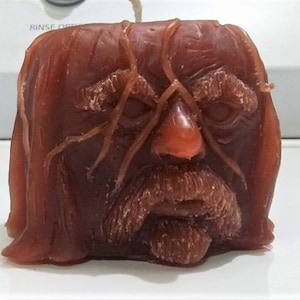 May include: A brown wax candle shaped like a human face with a long beard and a furrowed brow. The candle has a wick sticking out of the top.