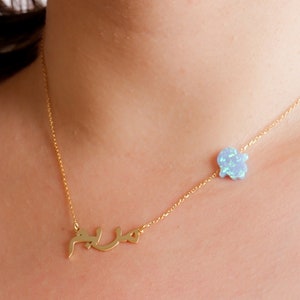 May include: A gold-tone chain necklace featuring a script nameplate pendant in Arabic calligraphy and a small, light blue, four-petaled flower charm positioned asymmetrically along the chain, resting against a plain white fabric background.