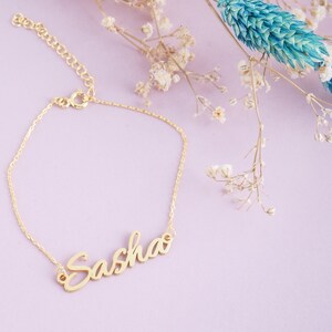 May include: A gold-toned metal bracelet featuring a delicate chain and a central nameplate with the name Sasha written in a cursive script font. The bracelet includes a small circular clasp and an adjustable chain link extension at the end.