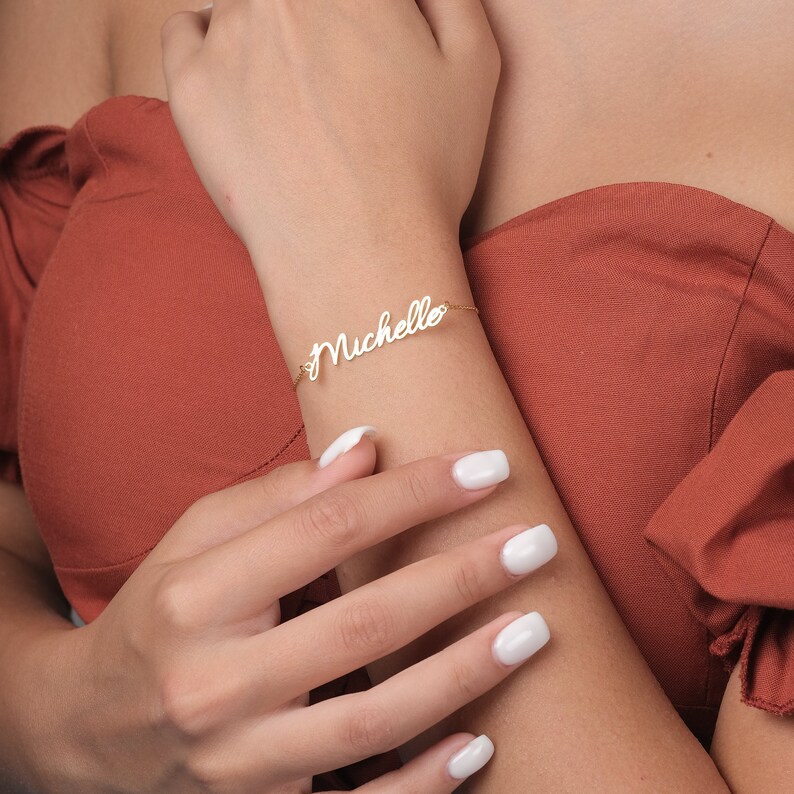 May include: A gold-toned metal bracelet featuring the name Michelle in a cursive script font. The nameplate is attached to a delicate chain that wraps around the wrist. The metal has a polished finish that reflects light, creating a bright appearance.