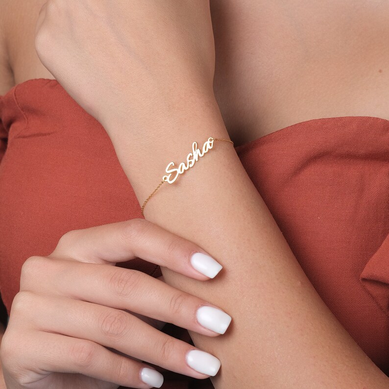 May include: A gold-toned chain bracelet featuring a central nameplate with the name Sasha written in a cursive script font. The delicate metal chain connects to both sides of the nameplate, creating a continuous piece of jewelry for the wrist.
