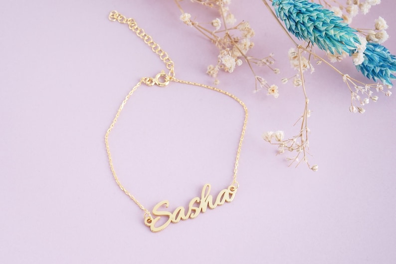 May include: A gold-toned metal bracelet featuring a delicate chain and a central nameplate with the name Sasha written in a cursive script font. The bracelet includes a small circular clasp and an adjustable chain link extension at the end.