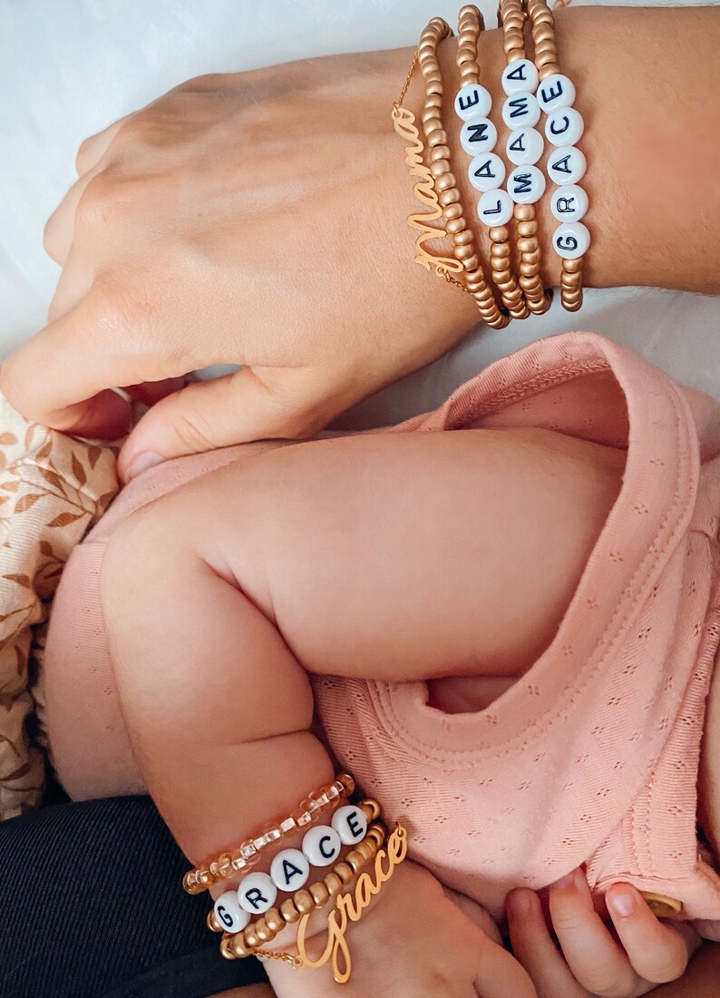 May include: A set of gold-toned beaded bracelets featuring white letter beads spelling out MAMA, LANE, and GRACE, alongside gold-toned script name charms. A matching set of smaller bracelets with white letter beads spelling GRACE is also visible.