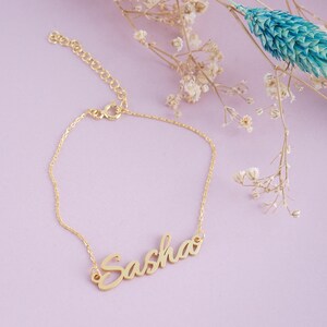 May include: A gold-toned metal bracelet featuring a delicate chain and a central nameplate with the name Sasha written in a cursive script font. The bracelet includes a small lobster claw clasp and an adjustable extender chain with circular links.