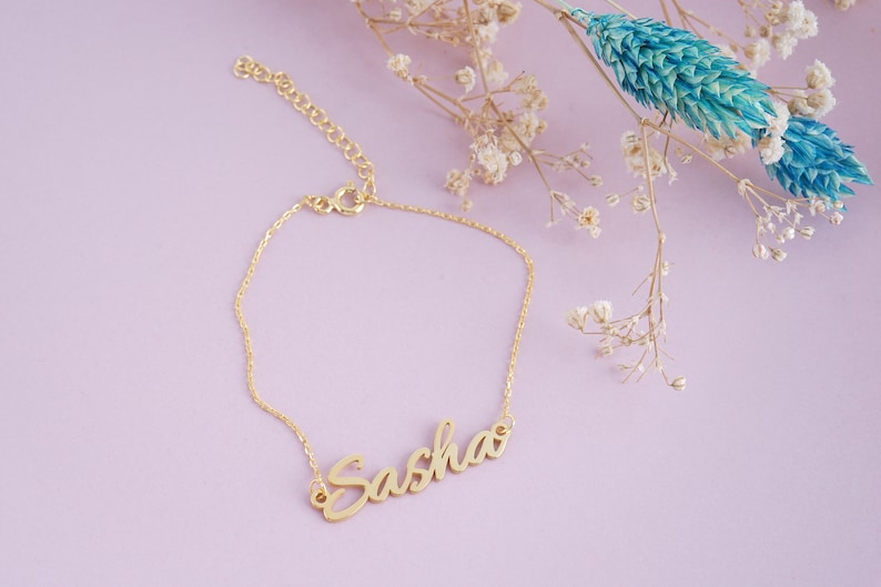 May include: A gold-toned metal bracelet featuring a delicate chain and a central nameplate with the name Sasha written in a cursive script font. The bracelet includes a small lobster claw clasp and an adjustable extender chain with circular links.