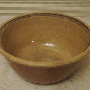 May include: A round, tan-colored ceramic mixing bowl with a smooth, glazed interior and a slightly textured, matte exterior surface. The bowl features a thick, reinforced rim and a deep, wide basin, showcasing a consistent earthy tone throughout.