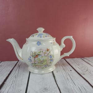 May include: A white teapot with a floral design and a gold trim. The teapot has a lid with a floral design and a handle on the side. The teapot is sitting on a white wooden surface.