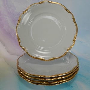 May include: A stack of four white porcelain plates with scalloped edges and a metallic gold trim. The plates feature a smooth, glossy finish and a curved, decorative border design that highlights the circular shape of each individual piece.