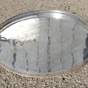 May include: A round silver-toned serving tray featuring a polished, reflective surface with an intricate, etched floral pattern in the center and a decorative, openwork lattice border along the raised outer rim of the metal platter.