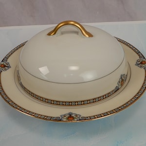 May include: A white porcelain butter dish with a domed lid and a gold handle. The dish sits on a matching plate with a decorative border featuring blue, orange, and gold accents. A classic serving piece.
