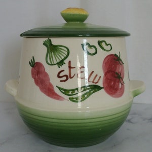 May include: Cream-colored ceramic stew pot with a green lid and base. The pot is decorated with hand-painted vegetables, including tomatoes, carrots, and onions, and the word "stew". The lid has a yellow and green knob and the pot has two handles.