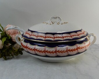 May include: A white porcelain tureen with a lid and two side handles, featuring a deep blue band decorated with a repeating orange and pink leaf pattern, accented with gold trim along the edges and a decorative handle on the lid.