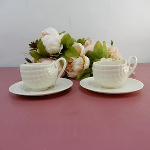 May include: Two white teacups and saucers with a textured basketweave design. The cups have curved handles and sit on matching saucers. A bouquet of light pink and white flowers with green leaves is in the background.