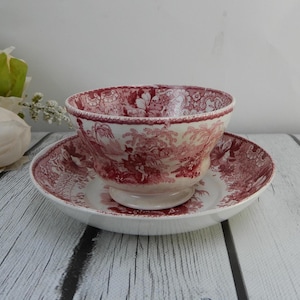 May include: A red and white ceramic bowl and saucer set. The bowl is round with a pedestal base, and the saucer is flat. Both pieces feature a detailed, repeating pattern of a landscape scene. The set is placed on a wooden surface.
