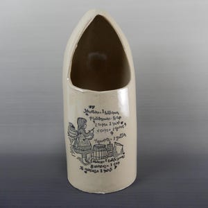 May include: Off-white ceramic utensil holder with a wide opening and a rounded top. The holder features a black illustration of a girl and text with measurements. A charming addition to any kitchen.