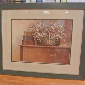 May include: A framed print featuring a still life scene of a woven basket filled with white baby's breath flowers, stacked round wooden boxes, and a wooden cabinet. The artwork is enclosed in a dark gray frame with a wide, light-colored matting.