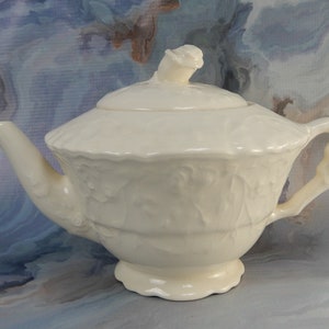 Puede incluir: Tetera de cerámica blanca con un pomo en forma de capullo de rosa en la tapa y un asa en forma de capullo de rosa. La tetera tiene un diseño floral en relieve.