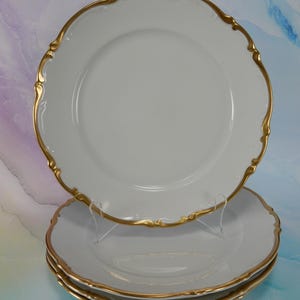 May include: A stack of white porcelain plates featuring a scalloped edge design with a metallic gold trim. The plates have a smooth, glossy finish and a circular shape, with one plate displayed upright on a clear stand while others are stacked below.