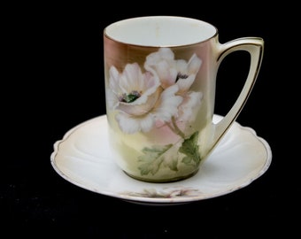 May include: A white porcelain teacup and saucer set with a floral design featuring pink and white flowers. The cup has a gold rim and a handle.