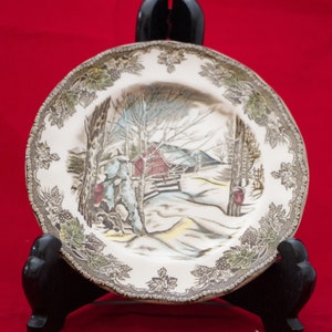 May include: A vintage ceramic plate featuring a winter scene with a red barn, trees, and a person in a snowy landscape. The plate has a brown and green floral border.