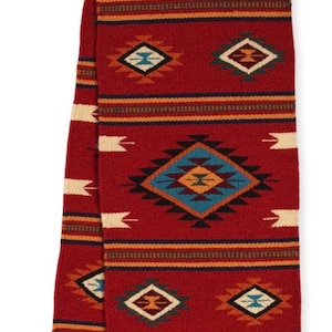 May include: A rectangular red scarf featuring a repeating geometric pattern in shades of blue, black, white, and orange. The design includes diamond shapes and horizontal stripes, finished with small red tassels at the bottom edges of the fabric.