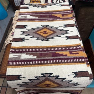 May include: A rectangular woven table runner featuring a cream-colored base with geometric patterns in shades of brown, maroon, and mustard yellow. The design includes repeating diamond motifs and horizontal stripes, finished with fringe on the ends.
