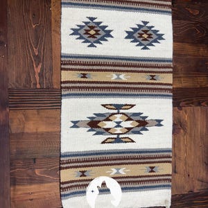 May include: A rectangular, woven table runner with a Southwestern design. The runner features a cream-colored background with geometric patterns in shades of blue, brown, and tan. The bottom of the runner has a white fringe. The text "COYOTE TRADERS RUSTIC FURNITURE" is at the bottom.
