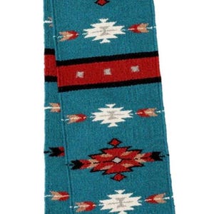 May include: A rectangular woven textile runner featuring a teal background with repeating geometric patterns in red, white, black, and tan. The design includes diamond shapes and horizontal stripes, finished with a gray border and small knotted tassels.