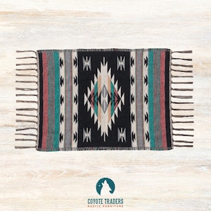May include: Rectangular woven rug with a black central design featuring geometric patterns in white, teal, and tan. The rug has vertical stripes in teal, gray, and red. It has fringed edges and the Coyote Traders logo.