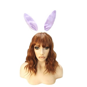 May include: A purple bunny ear headband with a white base. The headband is worn on a mannequin with long, wavy, auburn hair.