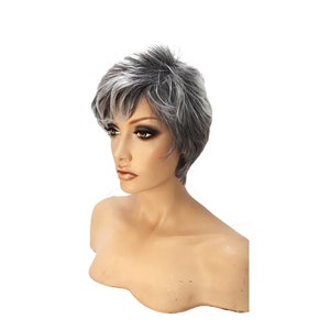 May include: A short, gray and white wig with a layered style. The wig is styled with a side part and has a natural, slightly tousled look.