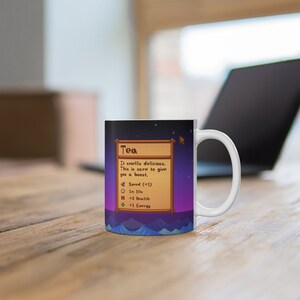 以下が含まれることがあります： 「Tea」というビデオゲームアイテムのピクセル化された画像が描かれたカラフルなデザインの白いセラミックマグカップ。マグカップには「It smells delicious. This is sure to give you a boost.」というテキストと、アイテムのステータス「Speed (+1), 1n 10s, +3 Health, +1 Energy」が記載されています。
