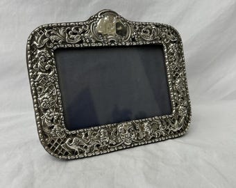 May include: A silver picture frame with an ornate design. The frame has a raised, textured border with floral and scrollwork details. The frame is empty and ready for a photo.