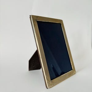 May include: A rectangular gold-colored picture frame with a dark blue interior. The frame has a textured border and decorative corner accents. A brown easel back supports the frame, which is angled slightly.