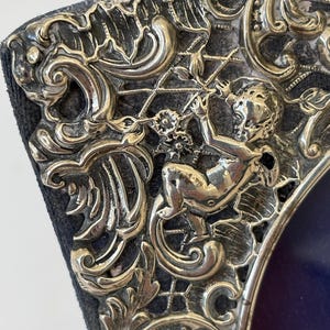 Puede incluir: Marco de fotos plateado ornamentado con un querubín detallado y un diseño de volutas. El marco tiene un respaldo azul oscuro y un borde decorativo. El marco es probablemente para una fotografía o una obra de arte.