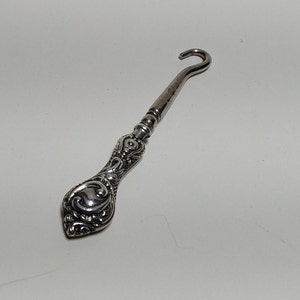 May include: A silver-toned shoe horn with an ornate, decorative handle. The handle is shaped like a teardrop and has a floral design.
