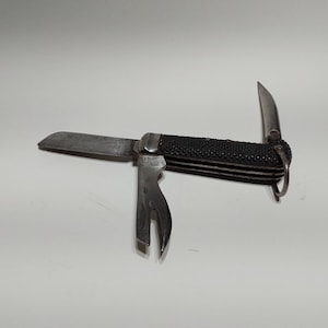 Puede incluir: Un cuchillo de bolsillo con mango negro, una hoja plateada, un abridor de botellas y una hoja pequeña. El cuchillo tiene un mango texturizado y un anillo en el extremo.