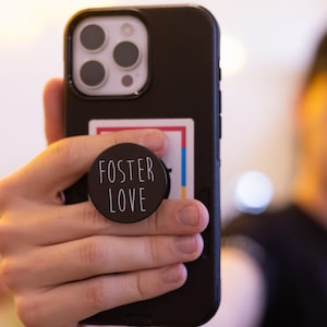 May include: A black smartphone in a black case with a round black pop socket that says "FOSTER LOVE" in white lettering. The phone is being held up in front of a blurred background.