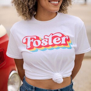May include: White t-shirt with the word "Foster" in red, outlined in white, with a retro wave design in yellow, blue, and red. The shirt is knotted at the waist and paired with blue jeans.