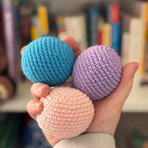 May include: Three small, crocheted balls in shades of blue, lavender, and peach are held in a hand. The balls have a textured surface and are spherical. The background is blurred, with a bookshelf visible.