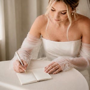 May include: A white strapless wedding dress with sheer, long, gathered tulle sleeves and a matching veil. The garment is worn while writing in a small, open, cream-colored notebook with a white pen, featuring a simple, clean aesthetic.