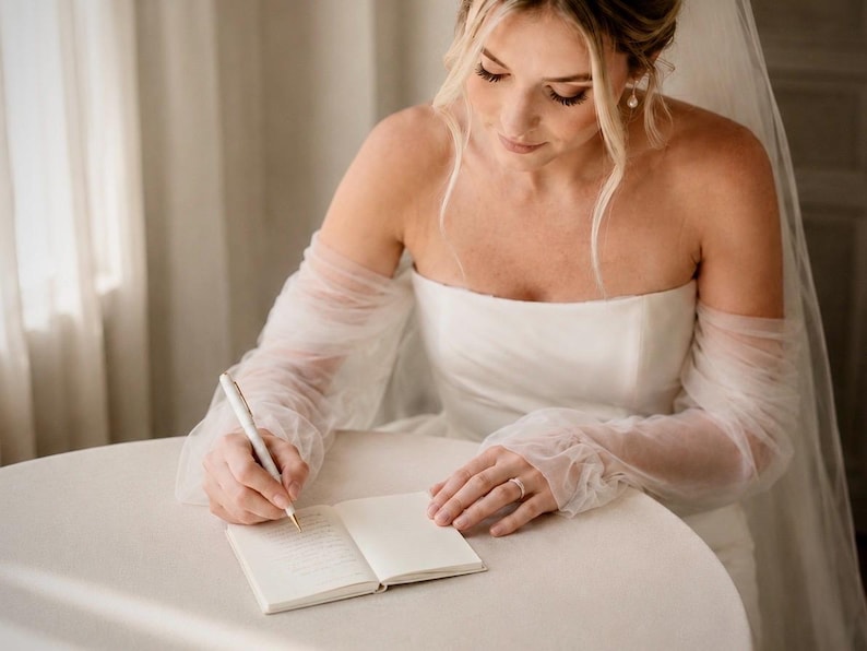 May include: A white strapless wedding dress with sheer, long, gathered tulle sleeves and a matching veil. The garment is worn while writing in a small, open, cream-colored notebook with a white pen, featuring a simple, clean aesthetic.