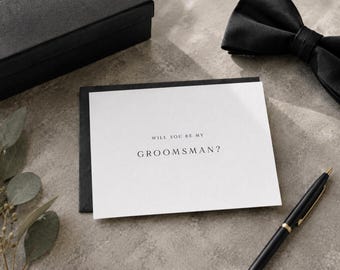 May include: A white rectangular card with black text that reads WILL YOU BE MY GROOMSMAN? The card is placed on top of a black envelope, accompanied by a black bow tie, a black pen, and a black box, all arranged on a textured grey surface.