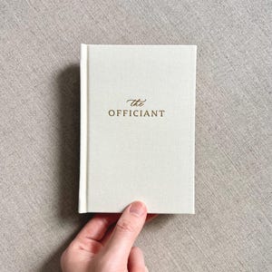 May include: A small, off-white book with the words "the Officiant" in gold lettering. The book is held by a hand against a neutral-toned fabric background. The book's cover has a textured appearance.